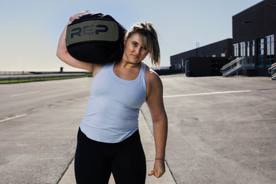 Woman holding REP stone sandbag on shoulder in an outdoor setting.